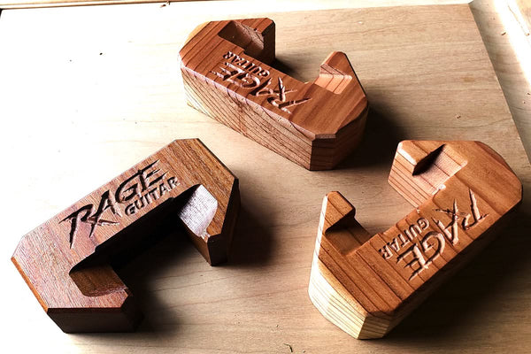 Three wooden guitar stands engraved with ROUTEK™ rest on a light surface. Their U-shaped design highlights the inscriptions, showcasing craftsmanship reminiscent of the precision associated with Hytek Tools CNC Industrial Router Machine.