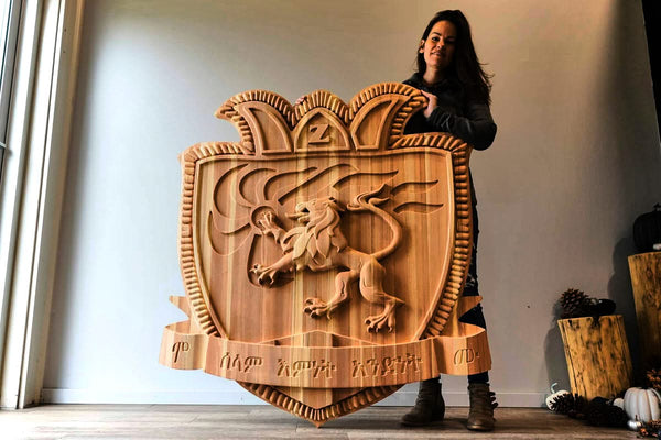 Indoors, a person holds an intricately carved wooden sign featuring a crowned lion emblem. The edges, crafted using the Hytek Tools 2024 ROUTEK™ CNC Industrial Router Machine Pkg, showcase decorative flair with text inscribed at the bottom.
