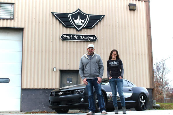 A man and a woman smile in front of a beige industrial building with Paul Jr. Designs signage. Nearby, a black car with custom designs gleams, showcasing precision stainless steel details crafted using the 2024 ROUTEK™ CNC Industrial Router by Hytek Tools under an overcast sky.