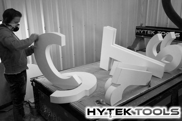 In a workshop, a person with a protective mask meticulously assembles large foam letters and numbers on a table. The space buzzes with machinery, including the impressive Aspire by Vectric - Version 11.5 CNC router, and Hytek Tools branding is visible in the background.