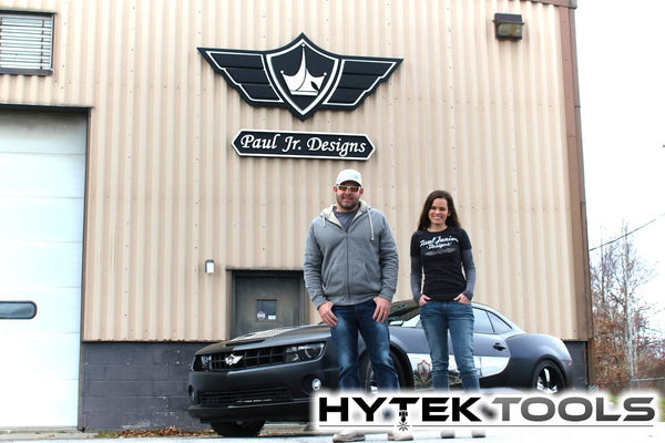 A man and woman stand by a black car outside a beige building with a Paul Jr. Designs sign, exuding a laid-back vibe. The building has a small garage door and displays the Hytek Tools logo, known for its Aspire by Vectric - Version 11.5 technology, at the bottom.