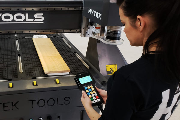 A person with long dark hair and wearing a black shirt operates a 2024 ROUTEK™ CNC Industrial Router Machine Pkg from Hytek Tools, guided by a handheld control device, with a wooden board on its precision rails.