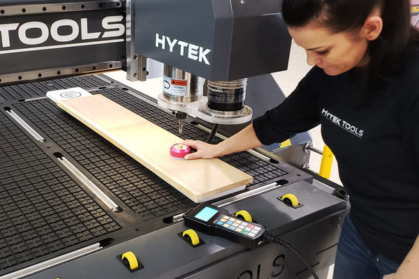 A person uses the 2024 ROUTEK™ CNC Industrial Router Machine Pkg to cut wood, holding a pink measuring tape on a wooden plank. The machine, by Hytek Tools, features precision rails and includes a visible control pad.