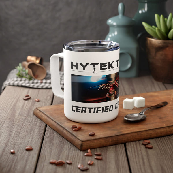 A white Insulated Coffee Mug by Printify with HYTEK and CERTIFIED printed on it rests on a wooden surface. Nearby are a spoon, sugar cubes, scattered coffee beans, a checkered cloth, and blue jars. A green plant complements the stainless steel design of this 10oz travel mug.