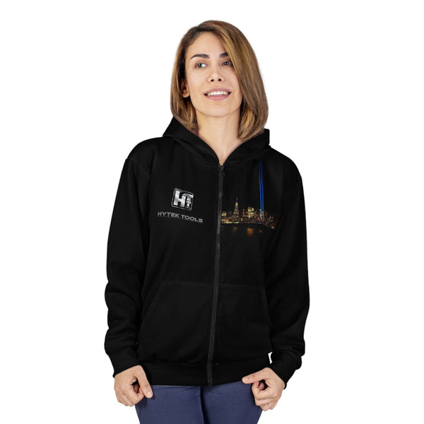 A person wearing a Printify Hytek Tools NYC Unisex Zip Hoodie featuring a cityscape, with shoulder-length hair, stands against a white background, smiling slightly.