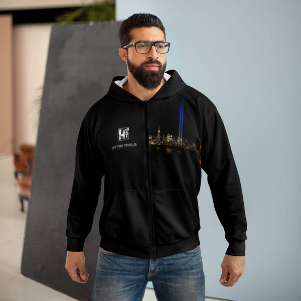A thoughtful man with a beard and glasses stands indoors, wearing the Printify Hytek Tools NYC Unisex Zip Hoodie featuring a cityscape design. One hand is in his pocket against a background of modern decor.