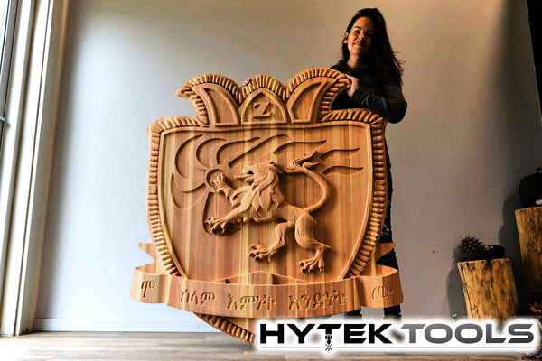 Indoors, a person holds a large, intricately carved wooden shield with a lion emblem, crafted with precision by the 2024 ATCTEK™ CNC Industrial Router machine. The designs and inscriptions are impeccable, featuring a HYTEK TOOLS logo at the bottom right.