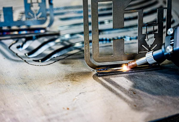 A close-up of a Hytek Tools WeldTEK™ RAYCUS Fiber Laser Welder 1000-3000w torch processing a metal sheet, emitting bright light while cutting or fusing intricate shapes. Similar patterned sheets are visible in the background.