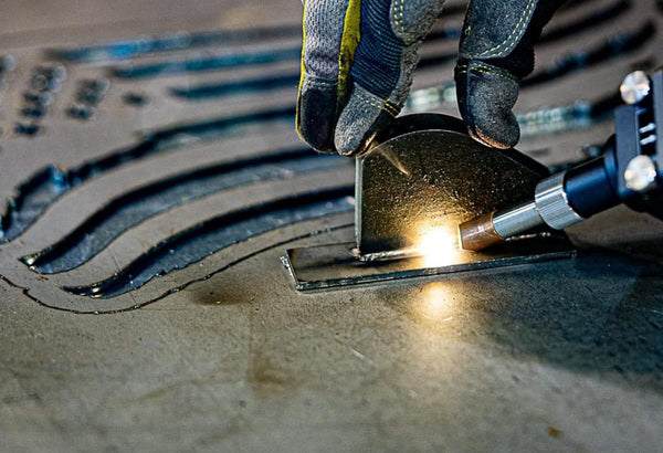A close-up captures a person wearing gloves using a plasma cutter on metal. Sparks fly from the bright arc, reminiscent of the WeldTEK™ RAYCUS Fiber Laser Welder 1000-3000w by Hytek Tools, as it slices through, revealing grooves from previous cuts.