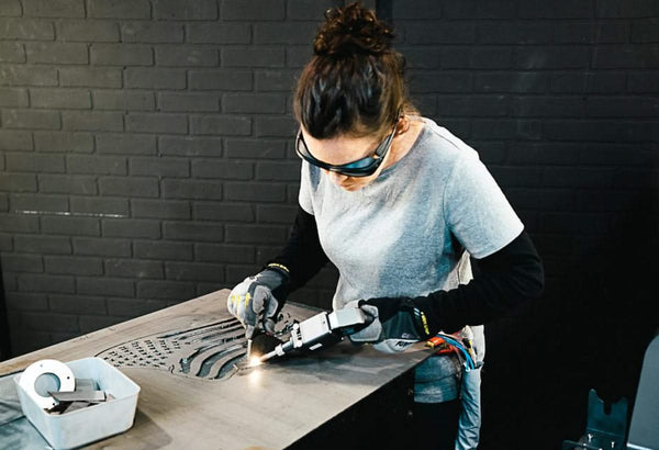 Wearing safety goggles and gloves, a person skillfully uses a handheld power tool to cut metal on a table. Nearby, the WeldTEK™ RAYCUS Fiber Laser Welder 1000-3000w by Hytek Tools hints at cutting-edge technology against a dark brick wall backdrop.